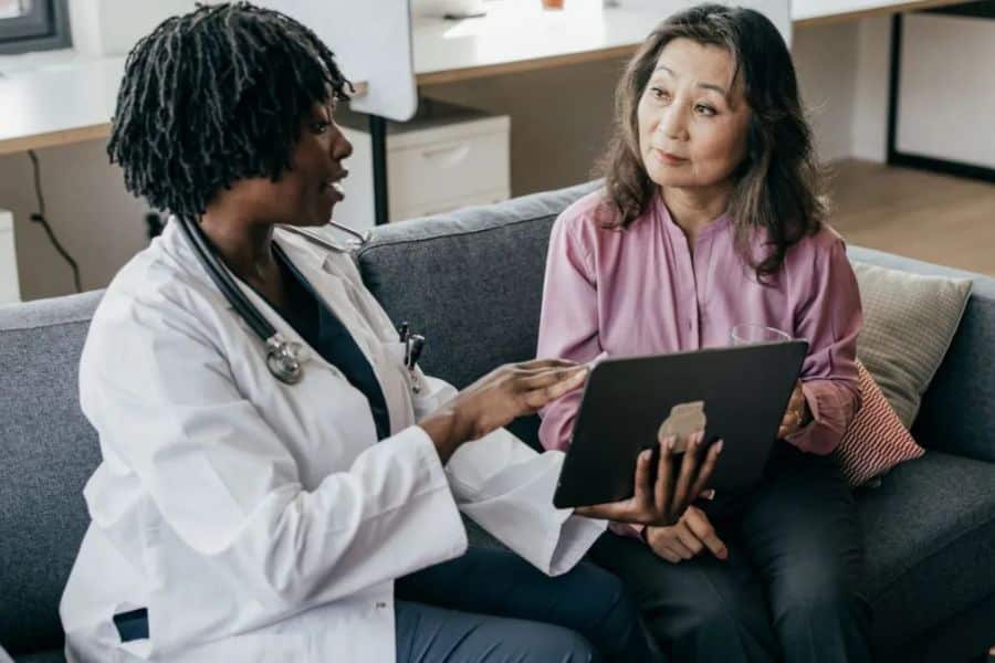 Doctor sitting on a couch with a patient and explaining information on a tablet. The visit shows a personal conversation about care and possible Medicare options.