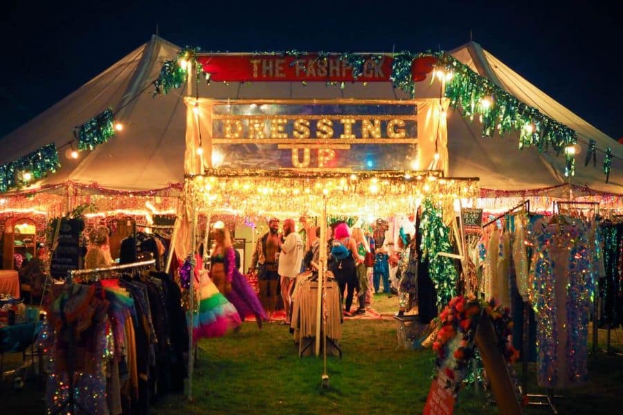 A brightly lit festival tent displays racks of colorful costumes and accessories at night. Signs read "THE FLASHBACK" and "DRESSING UP".
