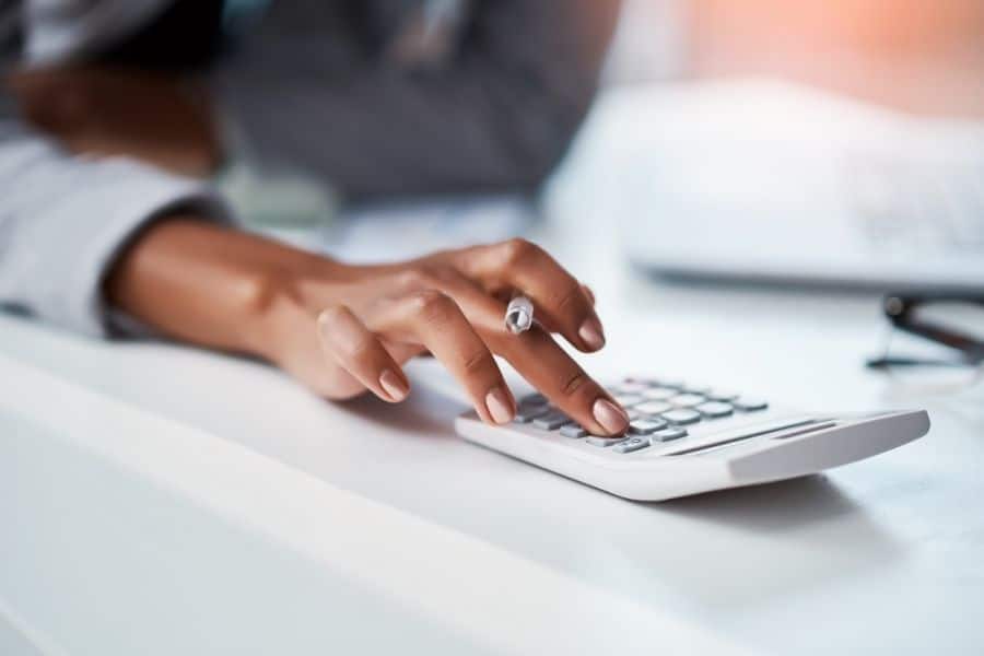 Close up of a person using a calculator at a desk while reviewing financial information. The image represents budgeting and financial calculations often involved in creating a catch-up plan for savings or debt repayment.
