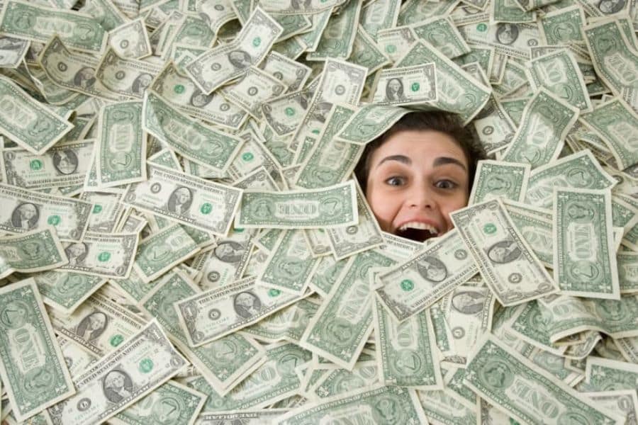 A smiling woman peeks out from a pile of scattered one dollar bills. The cash covers nearly the whole frame and gives the feeling of finding unexpected money.