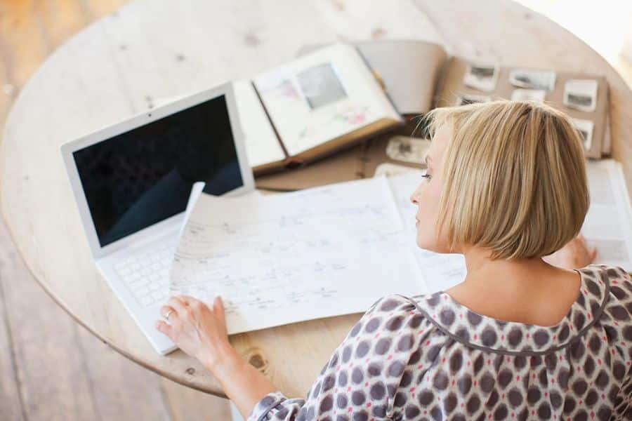 Woman working alone at a round wooden table with a laptop, notebooks, and planning documents spread around her. The workspace reflects organizing projects and managing multiple income streams from home.