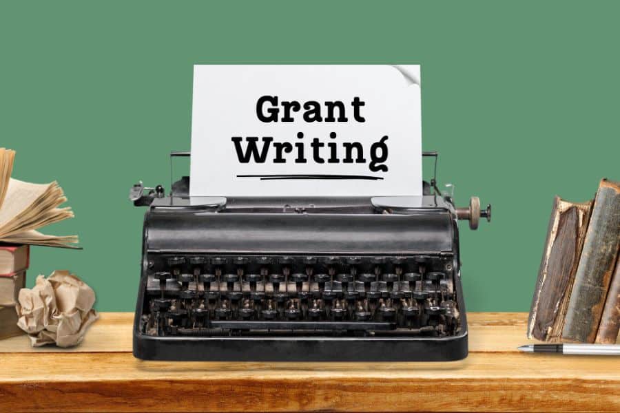 Vintage typewriter on a wooden desk with a sheet of paper that reads "Grant Writing" surrounded by books and papers. The image represents grant writing as a freelance income stream.