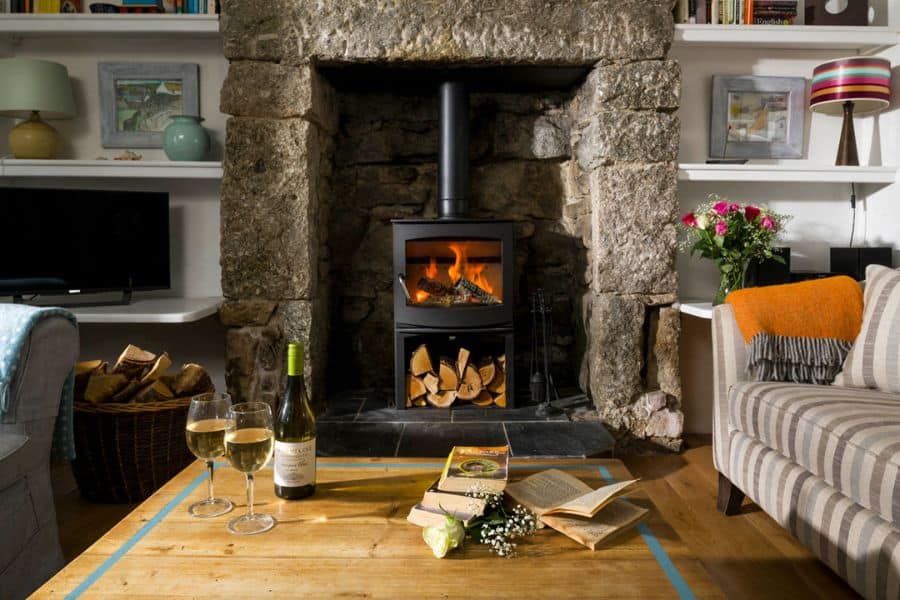 Cozy living room with a wood burning stove inside a stone fireplace, surrounded by bookshelves, blankets, and wooden furniture. The warm interior reflects sustainable switches focused on home heating efficiency and energy conscious living.
