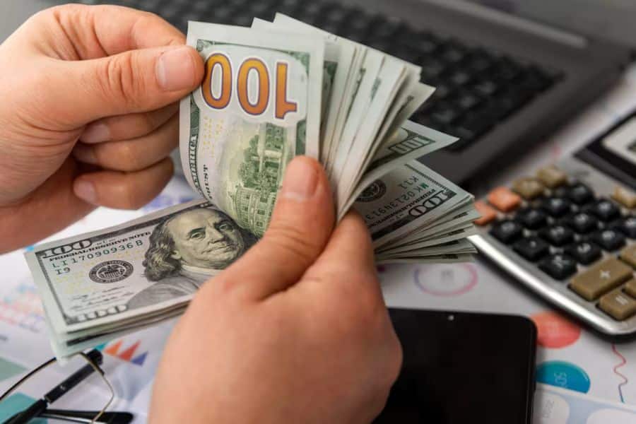 Close up of hands counting a stack of one hundred dollar bills beside a calculator, laptop keyboard, and financial paperwork on a desk. The scene represents budgeting, personal finance management, and building a savings account.
