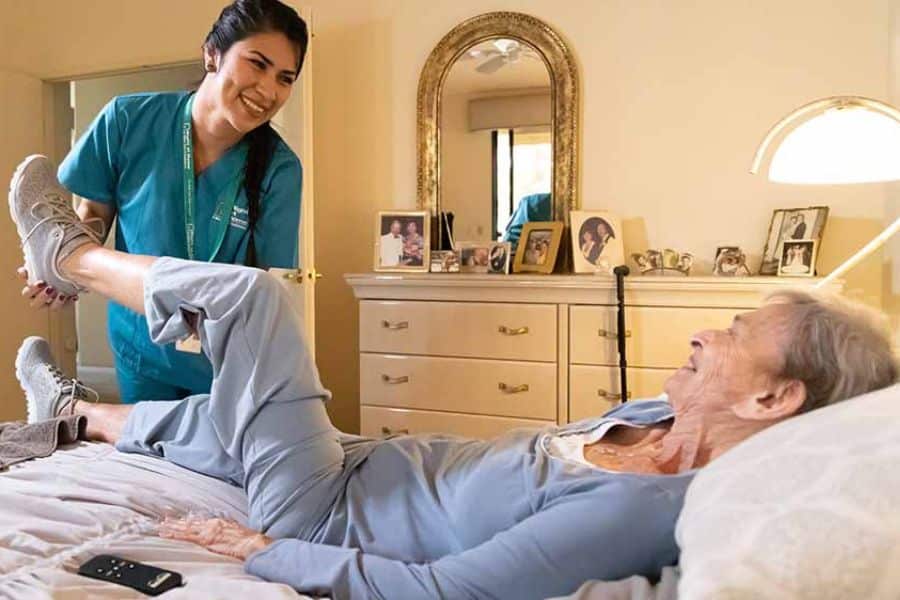 Caregiver in teal scrubs helping an older woman do leg exercises on a bed at home. The scene shows supportive recovery care in a bedroom.