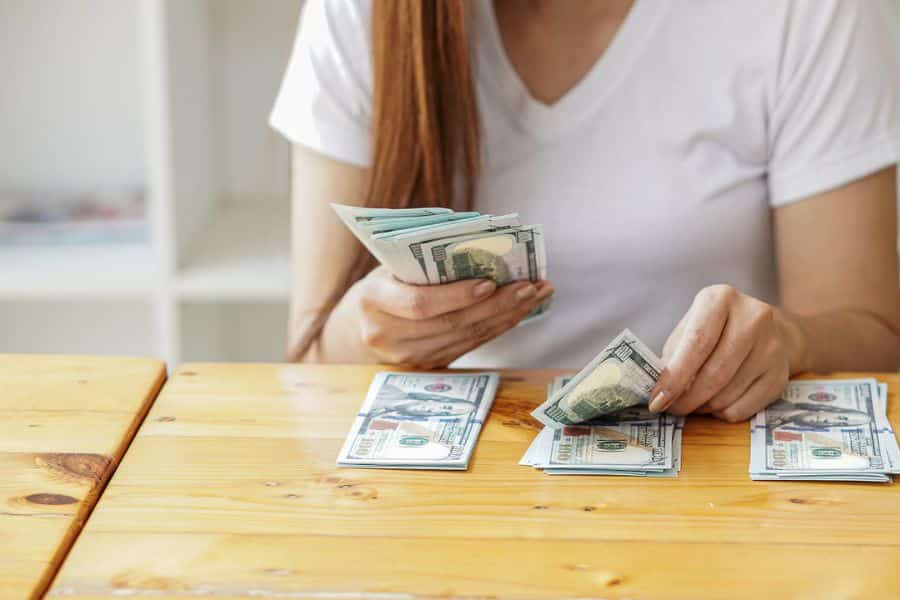 Woman sitting at a wooden table organizing and counting one hundred dollar bills into separate piles. The photo illustrates setting aside money for a savings account and managing household finances.