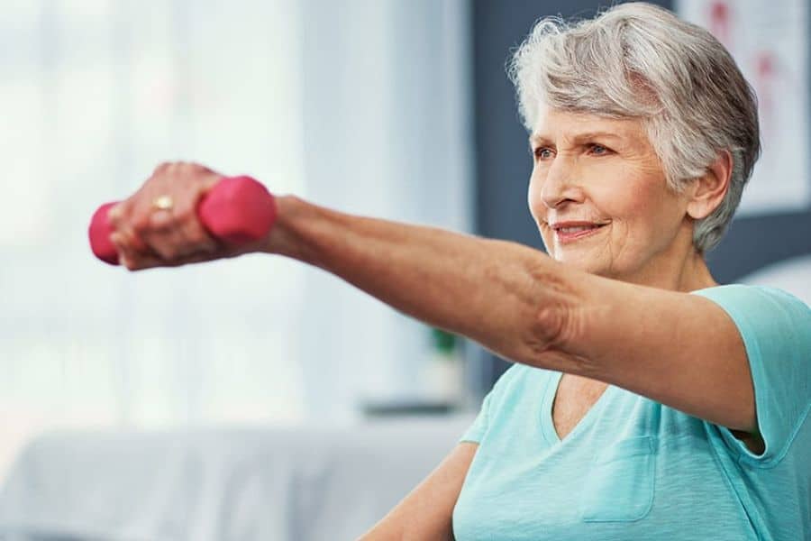 Older woman in a turquoise shirt doing a seated arm raise with a pink dumbbell during an exercise session.