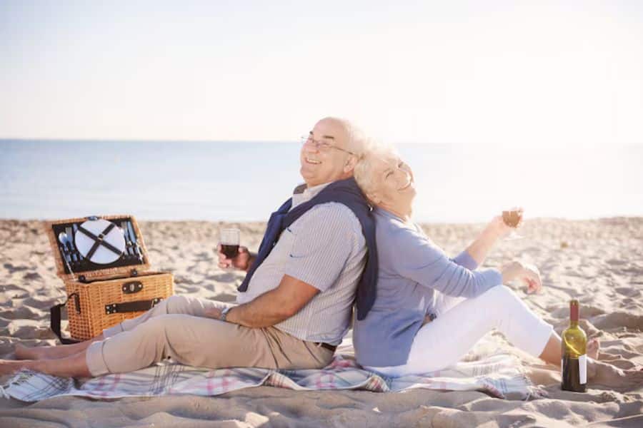 Retired couple sitting back to back on a beach blanket during a sunny picnic by the ocean with wine glasses and a picnic basket beside them. The relaxed setting reflects the lifestyle goals that can come from using the KonMari Method to simplify finances and prioritize meaningful retirement experiences.