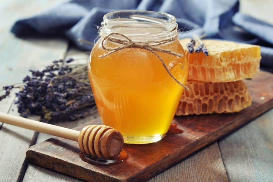 Glass jar of honey beside a honey dipper and pieces of honeycomb on a wooden table with dried lavender in the background. The natural ingredients and reusable glass container represent sustainable switches toward locally sourced and low waste food choices.
