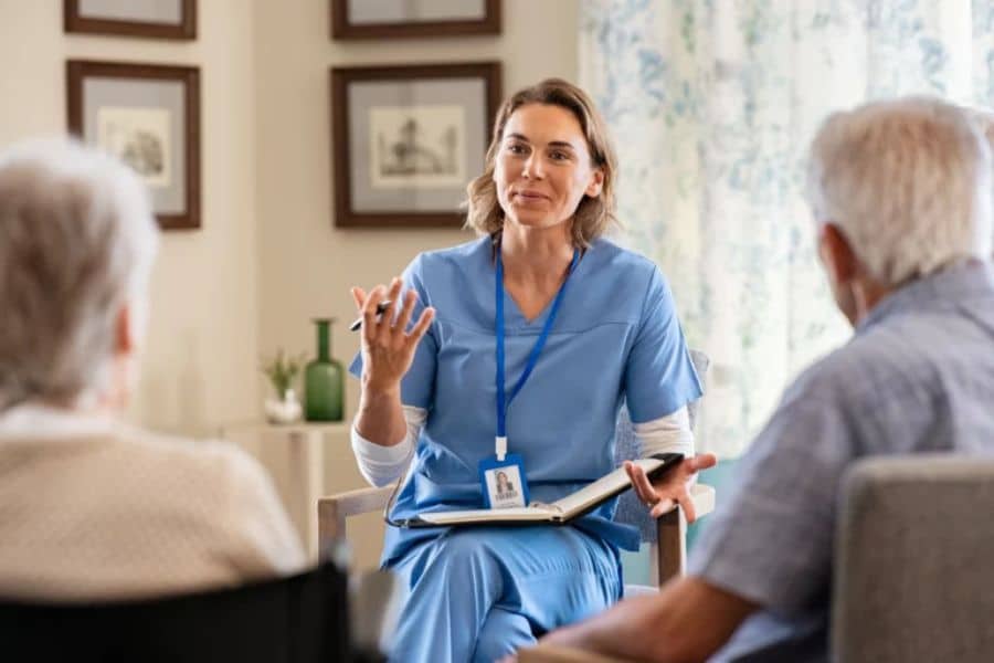 Nurse in blue scrubs talking with an older couple in a living room while holding a clipboard. The conversation appears focused on care planning and Medicare support.