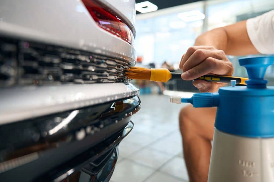 A person uses a brush and foam sprayer to clean the rear of a white car. The close up shows car detailing work that could become a simple side hustle.