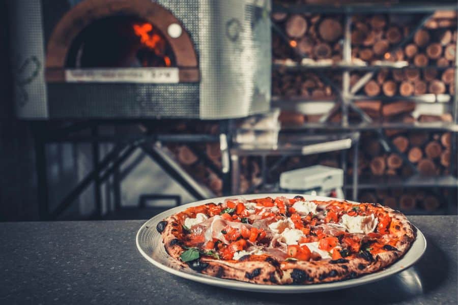 A fresh pizza sits on a plate in front of a wood fired oven and stacked logs. The restaurant scene highlights food service or catering work.