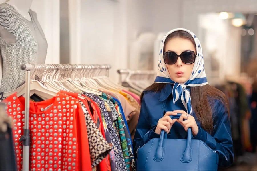 Stylish woman wearing sunglasses and a patterned scarf shopping beside a clothing rack filled with colorful garments. The photo suggests fashion resale or retail related income streams.