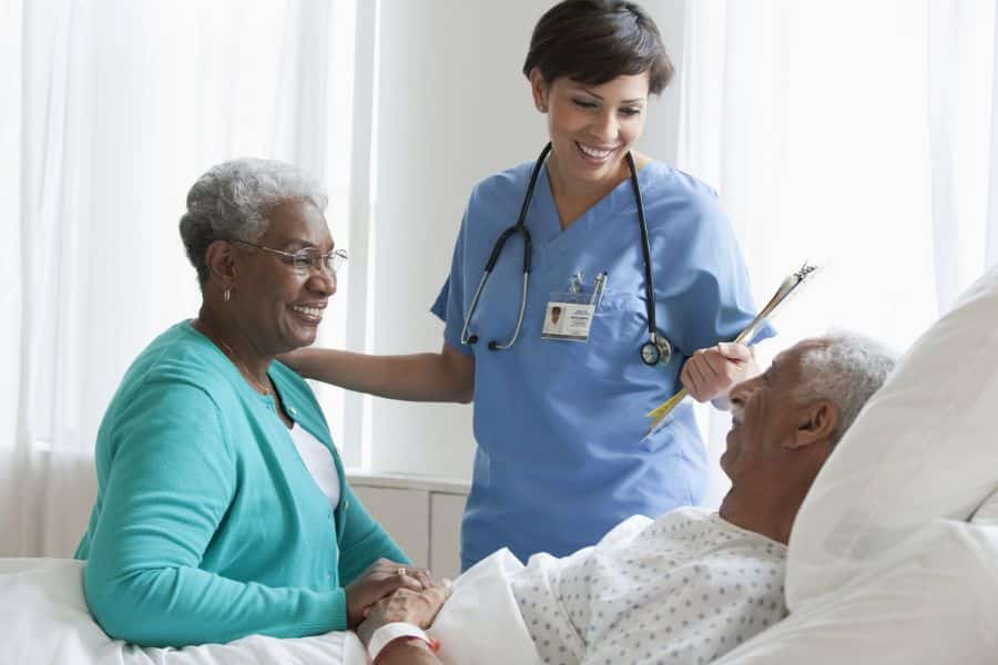 Nurse smiling with an older patient in a hospital bed and a visitor seated beside him. The scene shows coordinated care during a hospital visit.