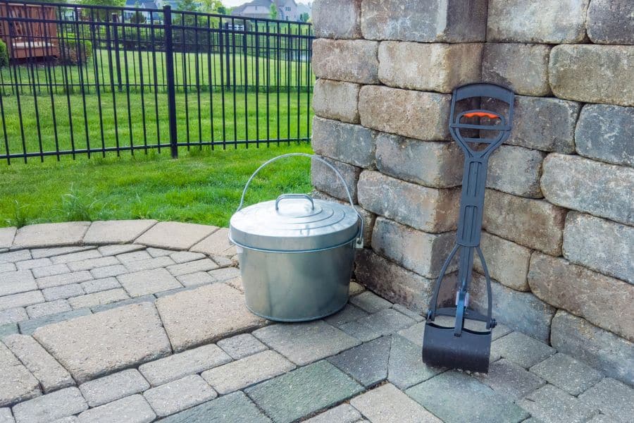 A metal ash bucket and long handled fireplace tool sit on a stone patio beside a retaining wall. The setup suggests outdoor cleaning or home maintenance work.