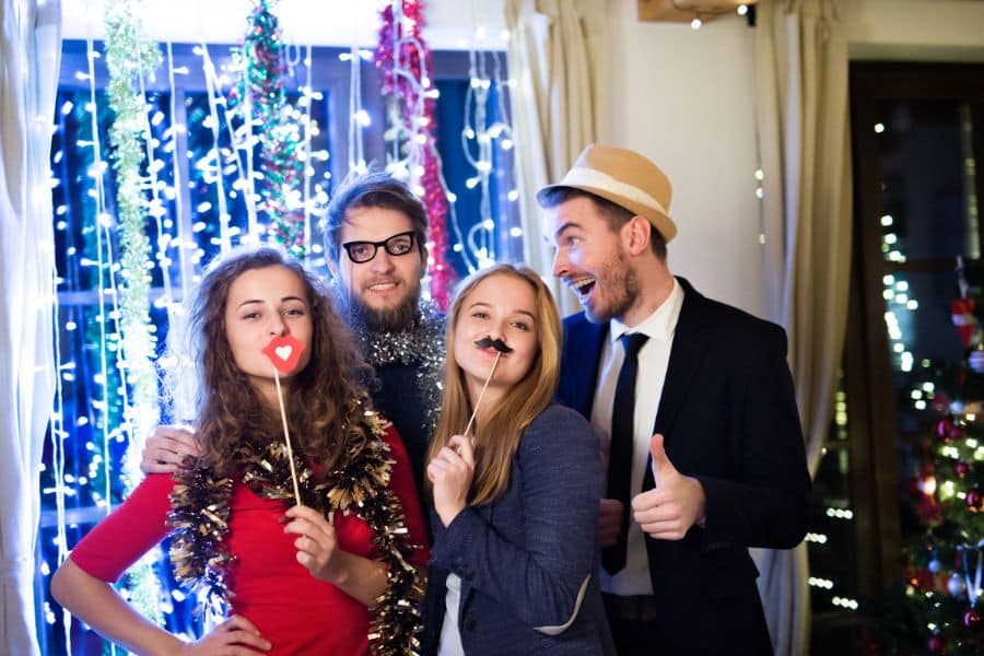 Four people pose with party props in front of bright holiday lights and tinsel. The festive scene could represent event work or party entertainment.