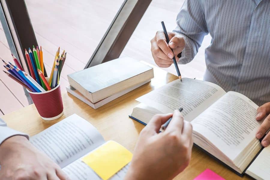 Two people studying open books together at a table with pencils, sticky notes, and colored pencils nearby. The scene represents tutoring, education, and academic income streams.