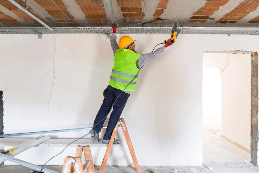 A construction worker in a hard hat and safety vest drills into a ceiling while standing on a ladder. The unfinished room shows hands on renovation work.