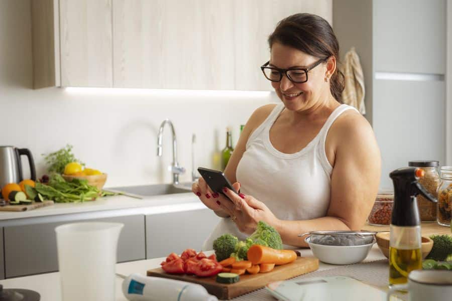 Woman smiling while checking her phone in a modern kitchen with chopped vegetables, broccoli, carrots, tomatoes, and cooking ingredients spread across the counter. The scene highlights healthy habits through meal preparation, home cooking, and intentional food choices.