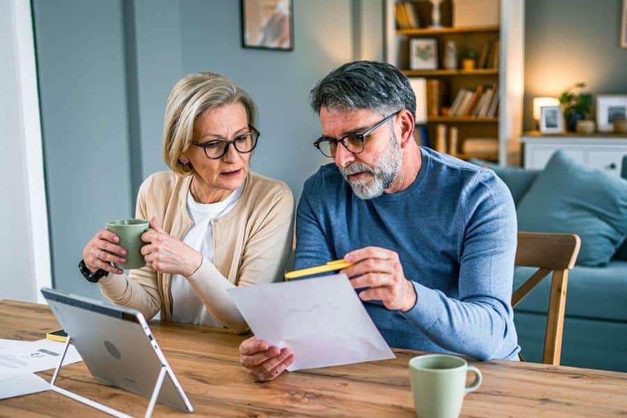 Older couple reviewing retirement fund paperwork together at a wooden table with a tablet and coffee mugs nearby. The man holds a pencil and document while the woman looks on with concern.