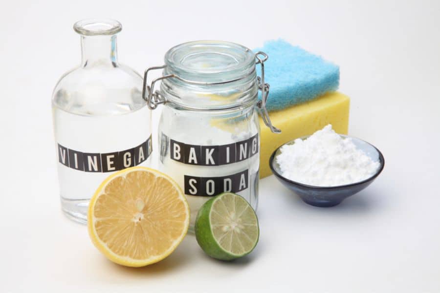 Bottle labeled "VINEGAR" beside a jar labeled "BAKING SODA" with lemons, limes, and cleaning sponges on a white background. The ingredients represent sustainable switches toward natural household cleaning products and reducing chemical cleaners.