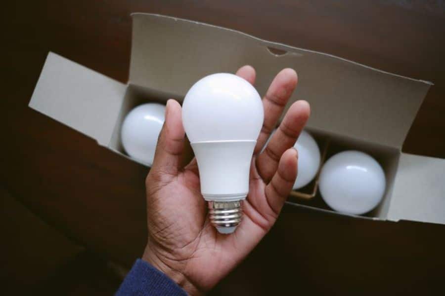 Hand holding an LED light bulb in front of an open box containing additional bulbs. The energy efficient bulb illustrates sustainable switches that reduce electricity use and household energy costs.