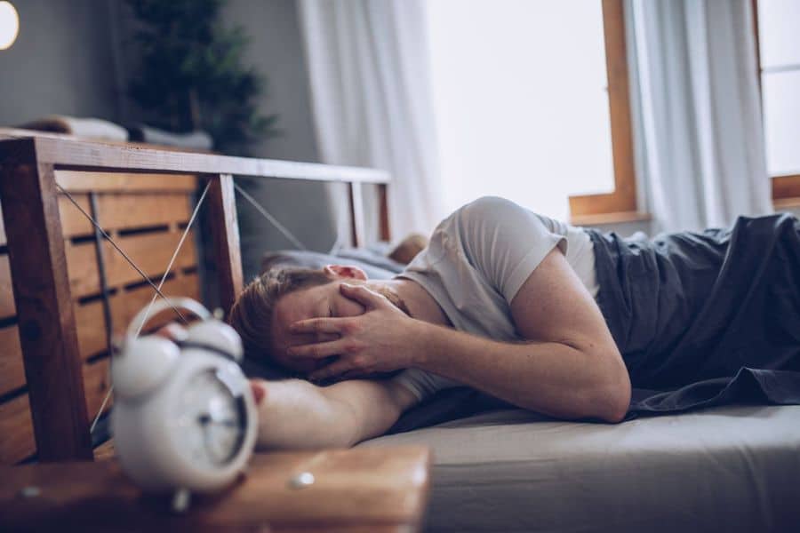 Man lying in bed with his hand over his face beside an alarm clock. The scene suggests restless sleep or waking up feeling tired.