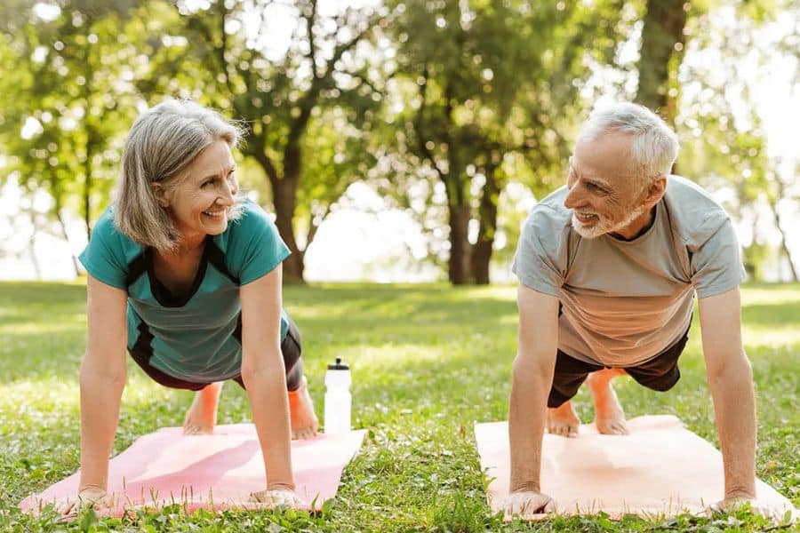 Older couple smiling at each other while holding plank poses on mats during outdoor exercise in a sunny park.