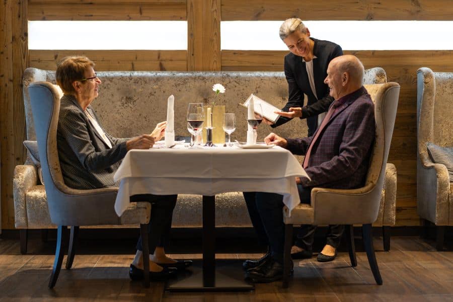 A server shows a menu to two older diners seated at a white tablecloth restaurant booth.
