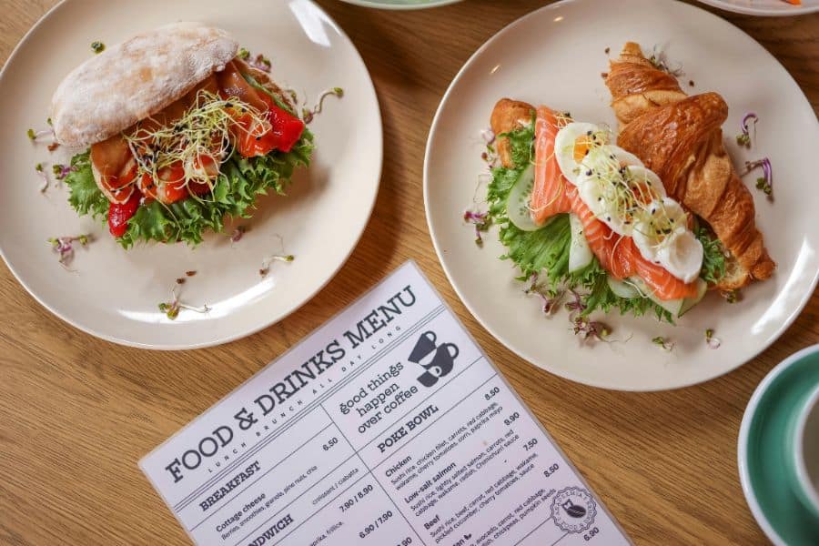 Two plated brunch sandwiches sit beside a printed food and drinks menu on a wooden table.