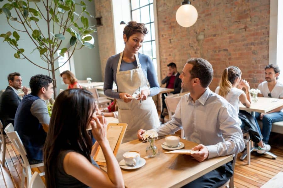 A café server takes an order from two diners holding menus at a busy restaurant table.
