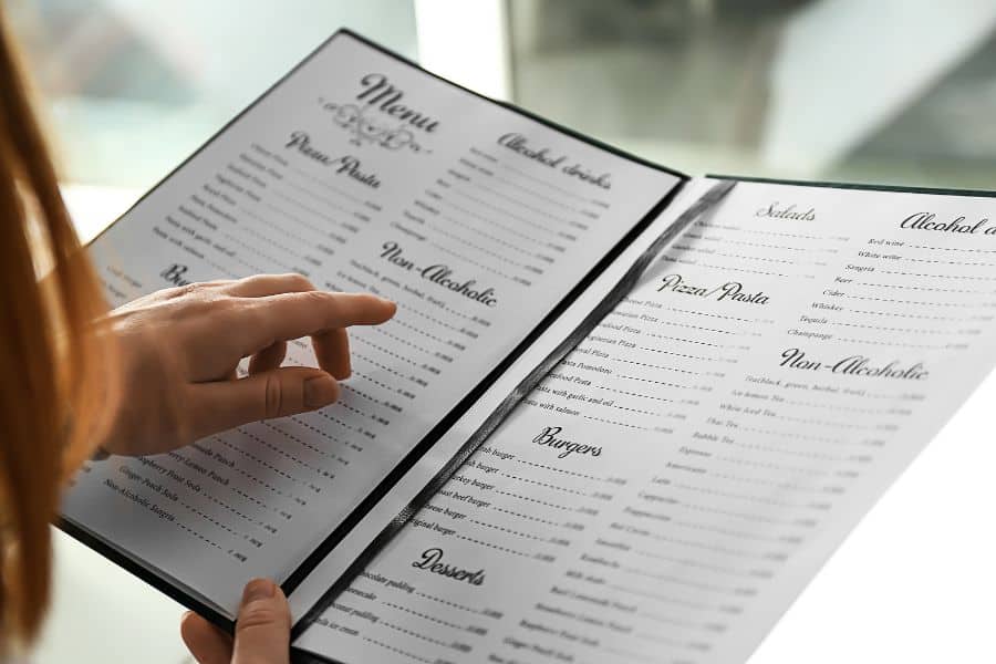 A person points at an open restaurant menu with sections for pasta. Salads. Pizza. Burgers. Desserts. Alcoholic drinks. Non alcoholic drinks.