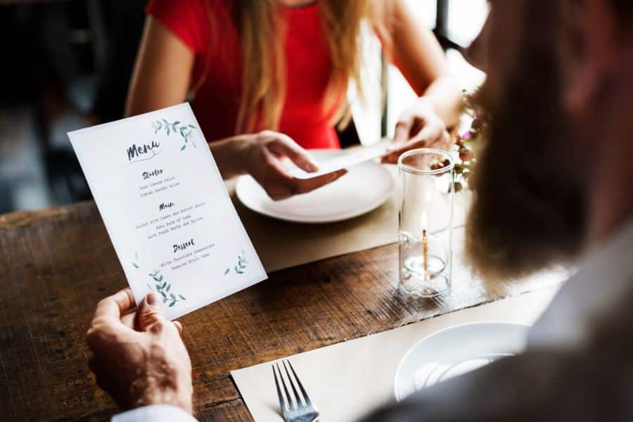 Diners look over a printed menu at a wooden restaurant table with plates and glasses.