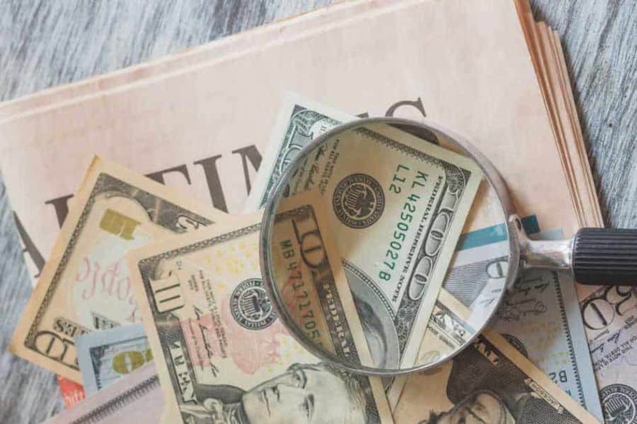 A magnifying glass enlarges part of a one hundred dollar bill sitting on top of papers and old currency. The close up suggests searching carefully through records to find missing or unclaimed money.