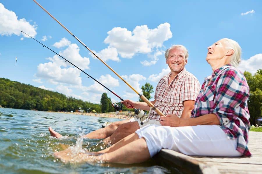 Happy older couple sits on a dock with fishing rods and their feet in the water on a sunny day. The relaxed outdoor scene highlights the type of leisure lifestyle many people hope to achieve through careful retirement rule planning.