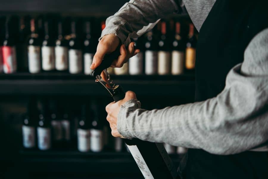 A restaurant worker opens a bottle of wine in front of shelves lined with bottles.