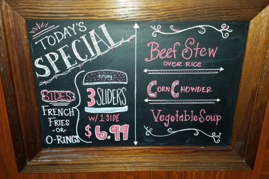 A restaurant chalkboard reads "Today’s special. 3 sliders w 1 side. $6.99. Sides. French fries or O rings. Beef stew over rice. Corn chowder. Vegetable soup."