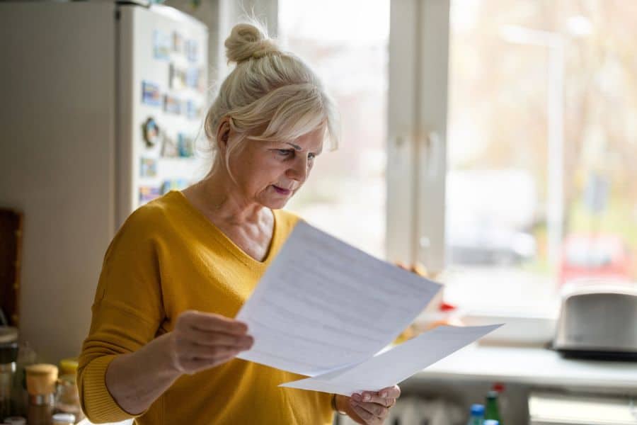 Older woman standing in her kitchen reading financial paperwork with a concerned expression near a bright window. The image represents the financial and tax planning challenges many surviving spouses face when dealing with the Widow’s tax.