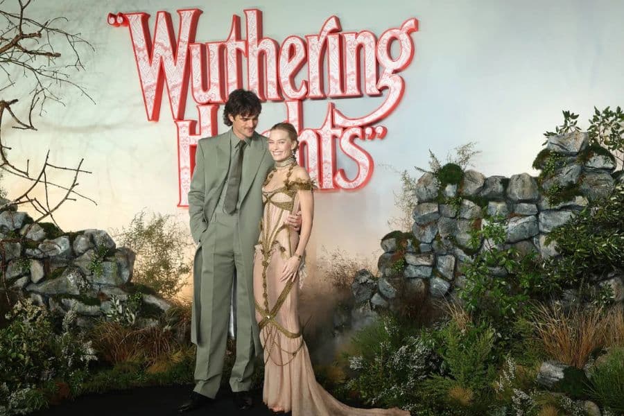 Two actors pose beside a glowing “Wuthering Heights” sign surrounded by stone ruins and dramatic greenery at a themed premiere event inspired by the gothic novel.