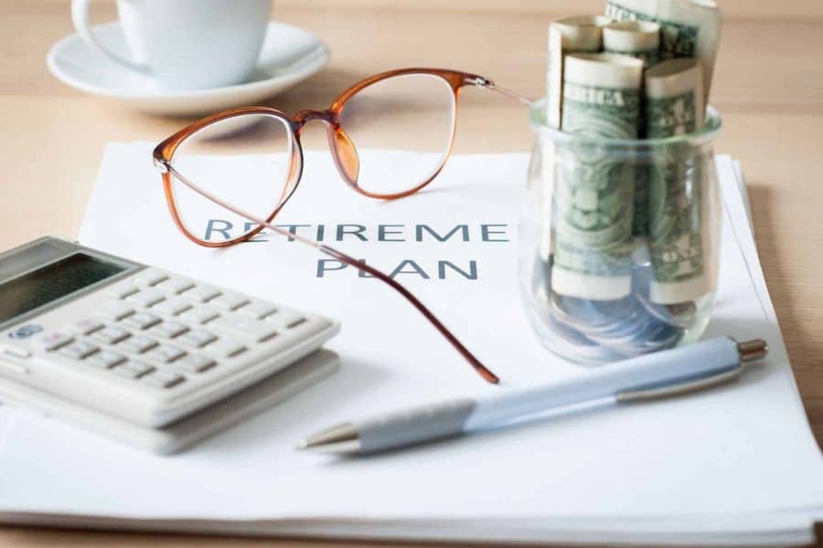 Retirement plan document on a desk beside a calculator, eyeglasses, rolled cash, and a pen. The arrangement represents long term financial planning and creating a catch-up plan for retirement savings.