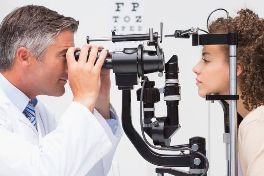Eye doctor performing an eye exam on a young woman using specialized ophthalmology equipment. The clinical vision screening may be part of preventive care or Medicare covered eye services.