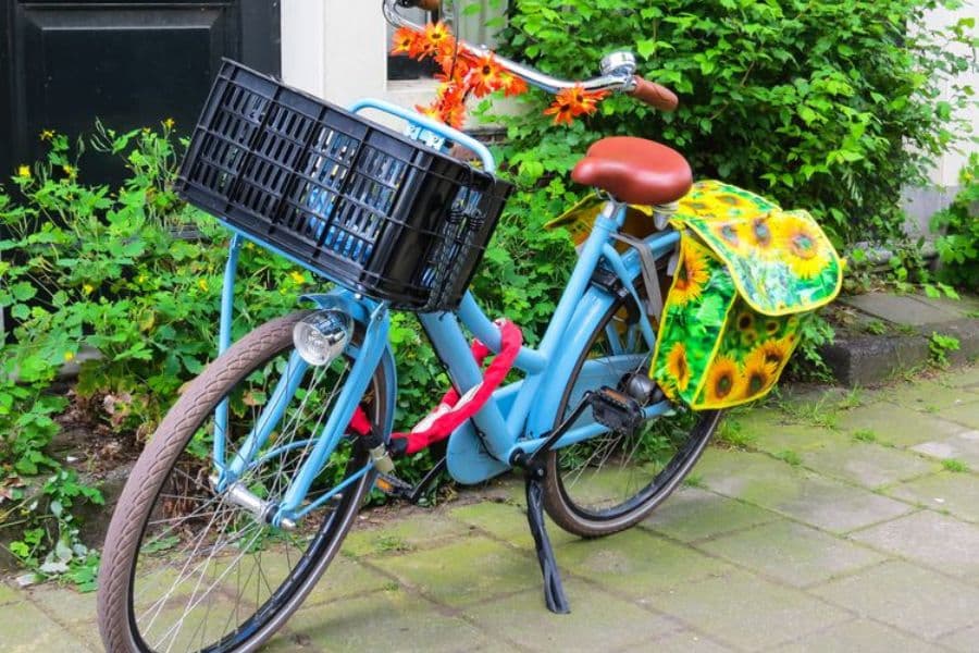 Blue bicycle with a front basket and sunflower patterned bags parked beside green plants on a sidewalk. The bike represents sustainable switches such as cycling instead of driving for short everyday trips.
