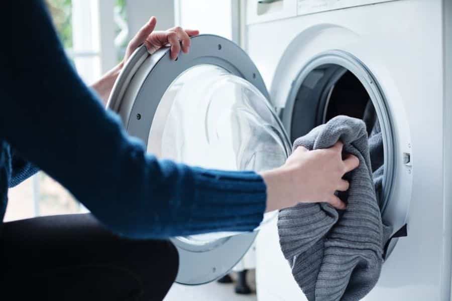 Person placing clothing into a front loading washing machine in a bright laundry room. The image reflects sustainable switches like energy efficient laundry habits and washing clothes only when needed.