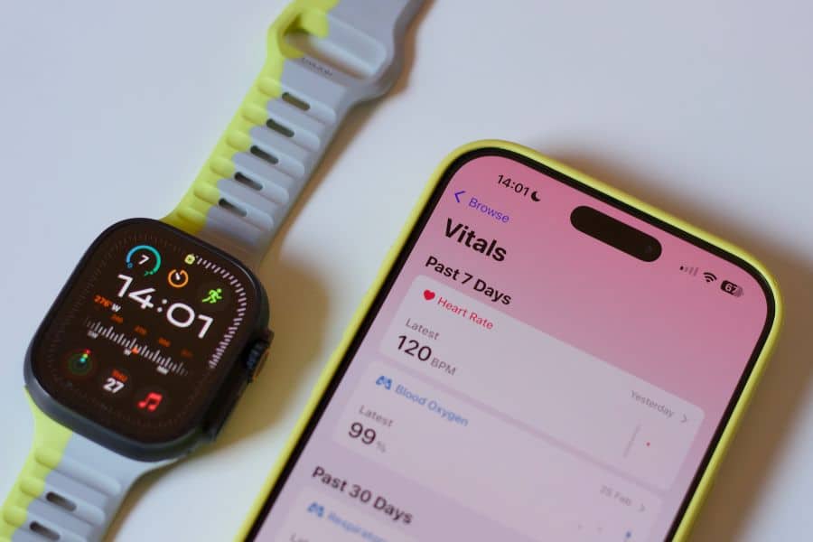 Apple Watch and iPhone displaying health metrics on a light surface. The phone screen reads "Vitals. Past 7 Days. Heart Rate. Latest. 120 BPM. Blood Oxygen. Latest. 99 percent" showing how a fitness app can track heart rate and oxygen data.