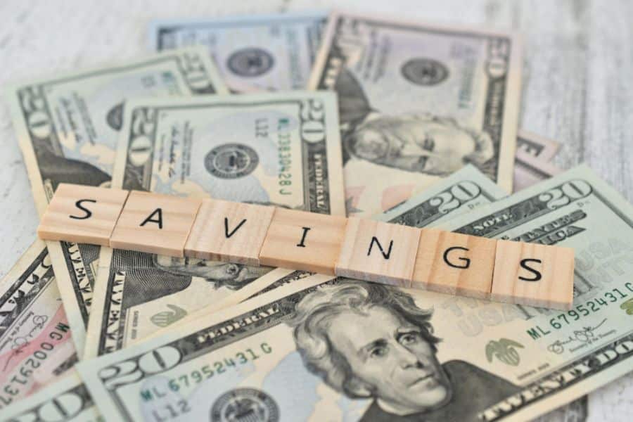 Wooden letter tiles spelling “SAVINGS” placed across scattered US dollar bills including twenties and fifties. The image highlights the concept of growing a savings account and building financial security.