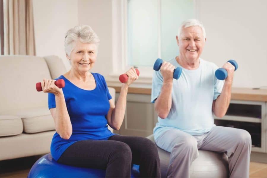 Older woman and man seated at home while lifting dumbbells for a light strength exercise routine.