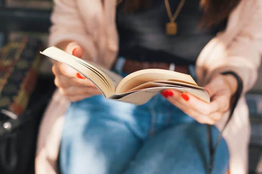 A seated woman with red painted nails holds an open paperback book in her lap. Reading offers a quiet way to engage the mind and strengthen concentration.