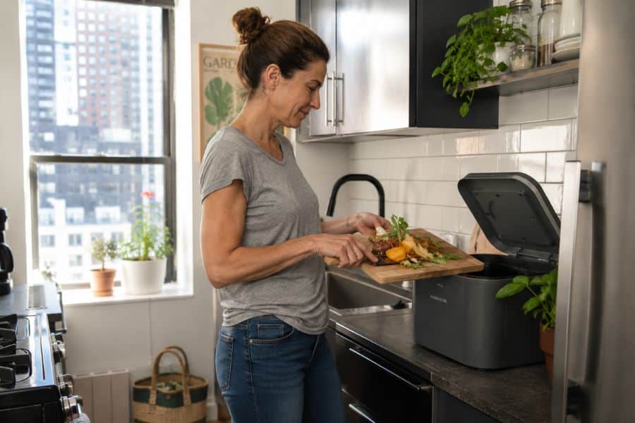 A woman scrapes vegetable peels and food scraps from a wooden cutting board into an electric kitchen compost bin. The bright apartment kitchen shows how composting can fit into everyday meal preparation.