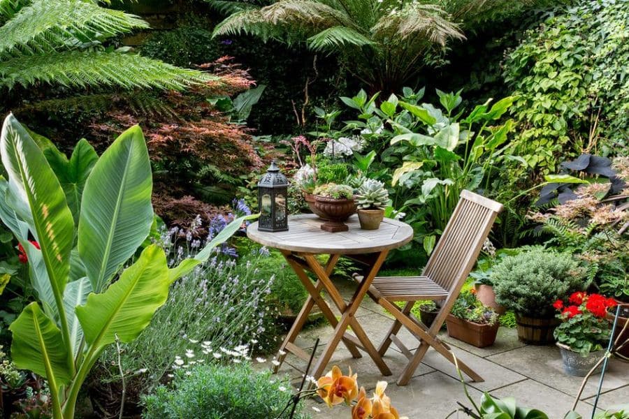 Tiny garden patio surrounded by lush tropical style plants, ferns, and flowering containers with a small table and chair set tucked into the greenery. The design shows how a tiny garden can create a private outdoor retreat in a limited space.