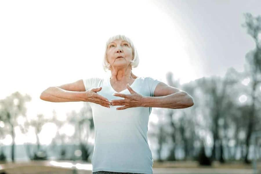 An older woman practices a slow mindful movement outdoors with her hands held in front of her chest. The gentle exercise suggests balance focus and calm concentration.